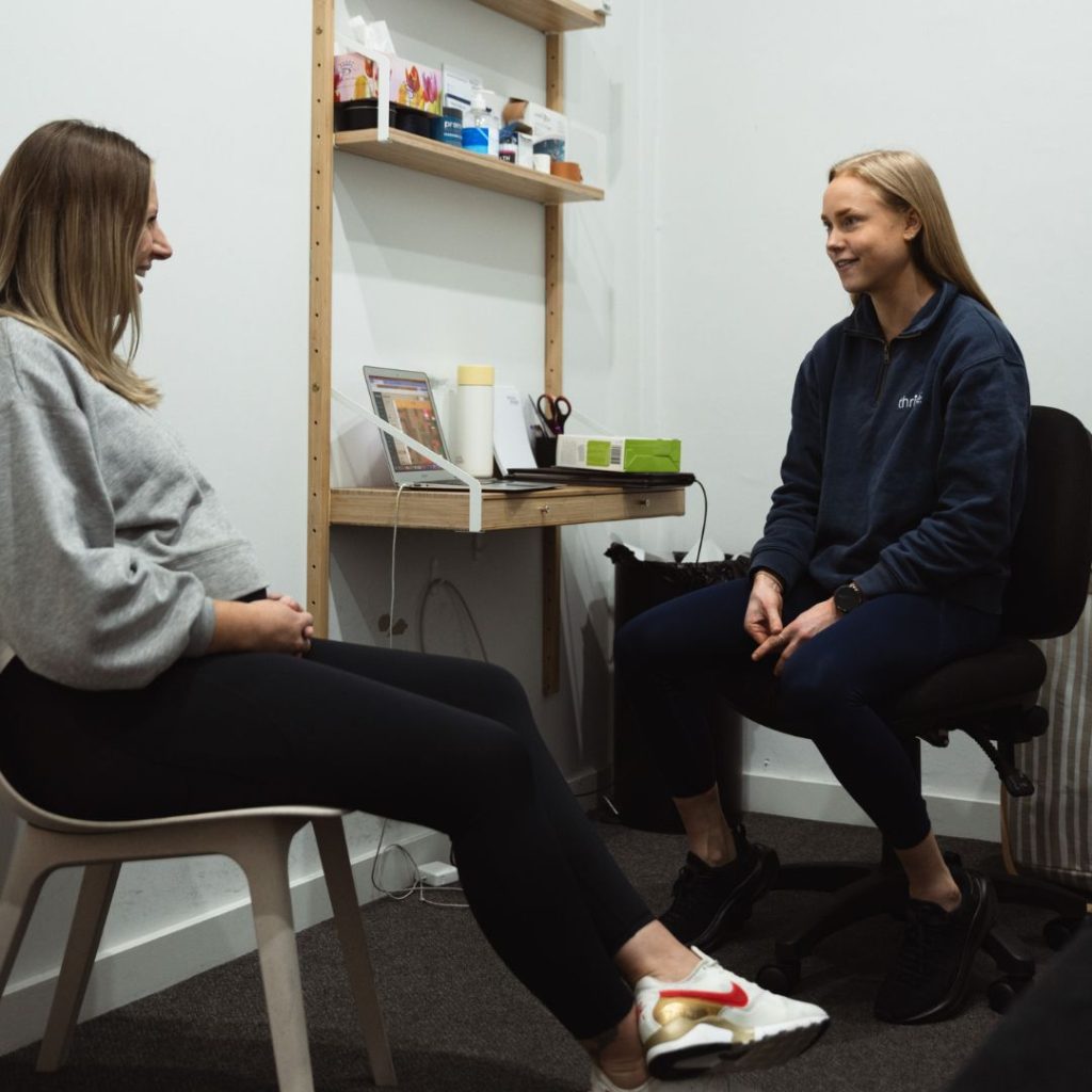 A physiotherapist holds a pelvic model while explaining pelvic floor anatomy to a seated woman during a consultation.